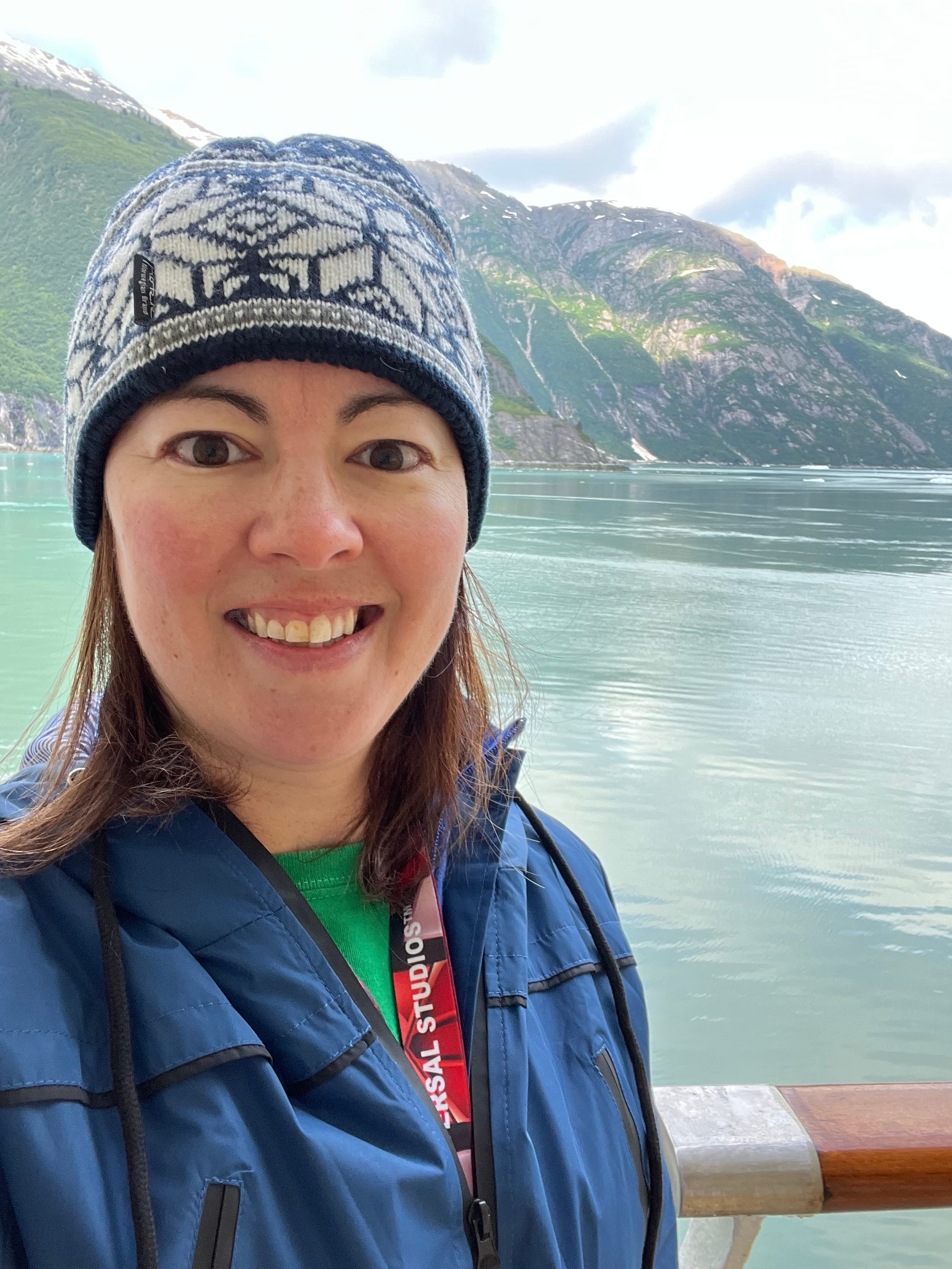Smiling woman in a blue jacket and knit beanie before scenic mountains and glacial water.