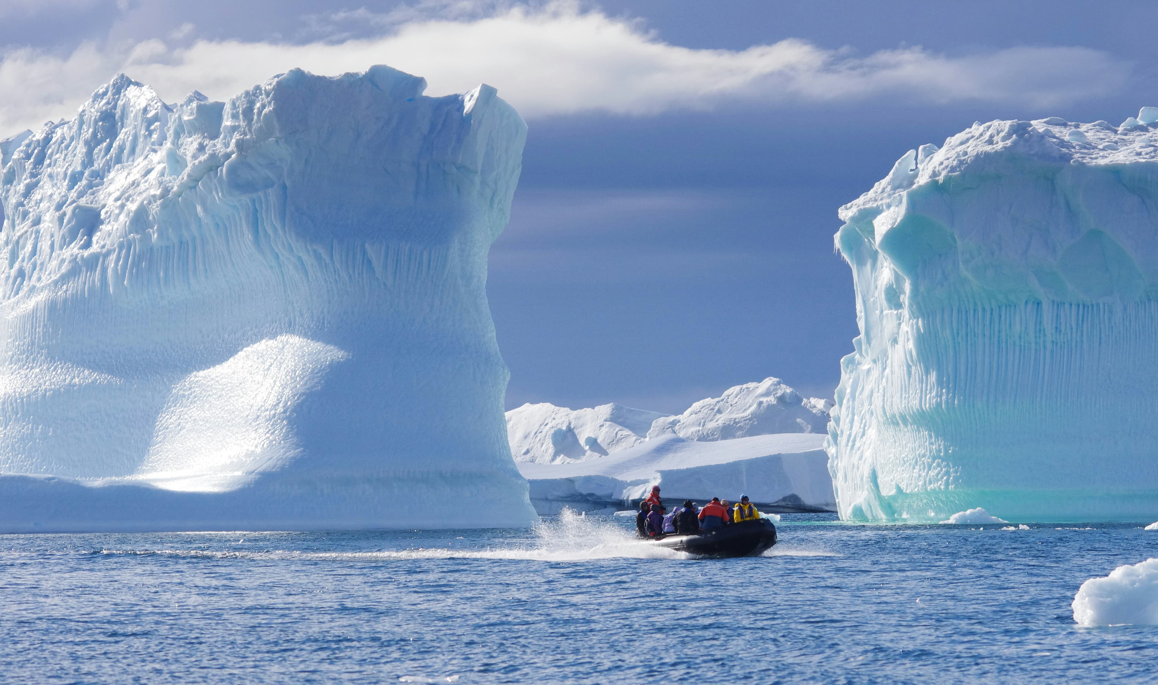 A black inflatable boat speeds through blue water between two massive, towering blue icebergs.