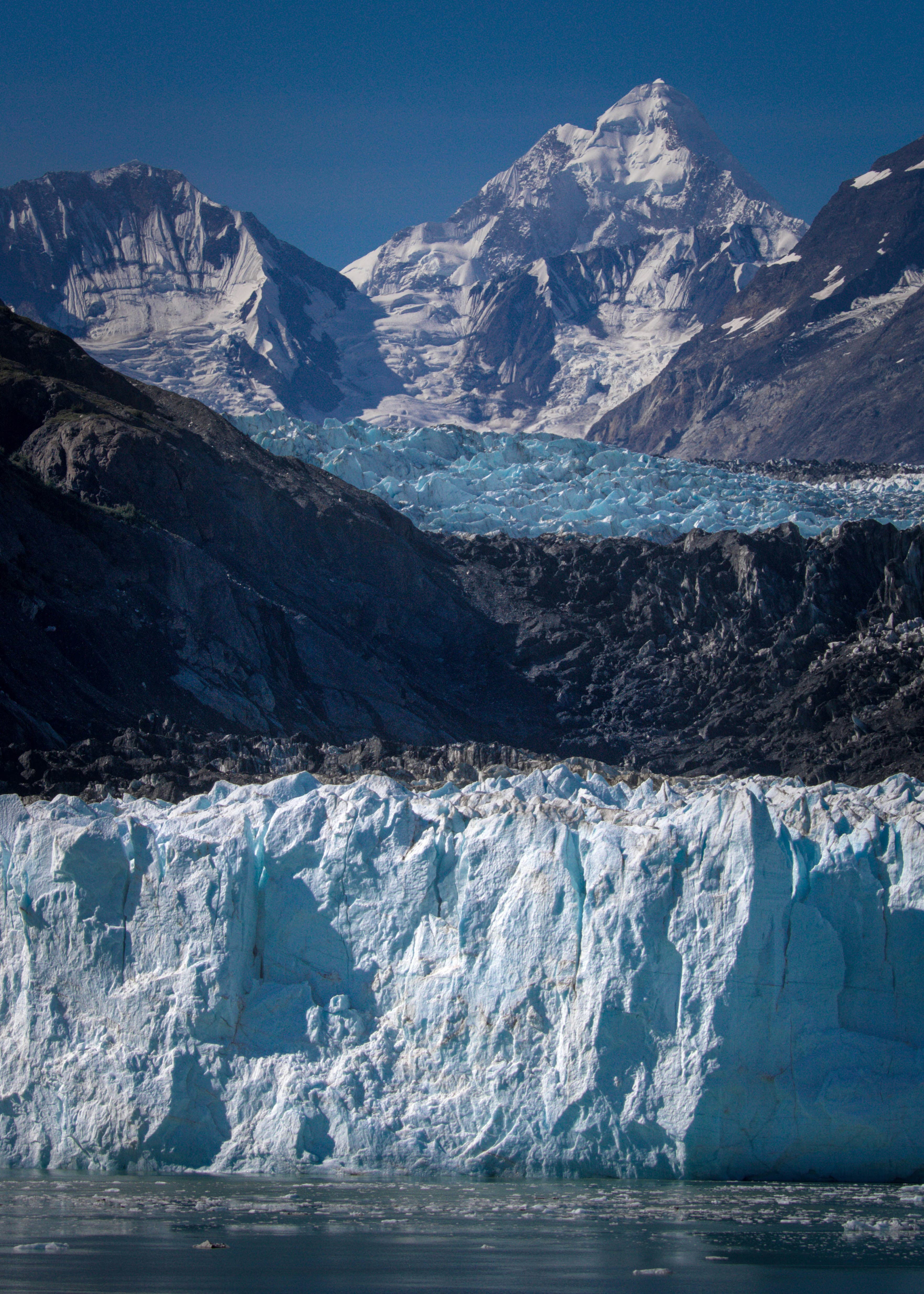Massive blue glacier wall stands before dark rocky mountains and a towering snow-capped peak.
