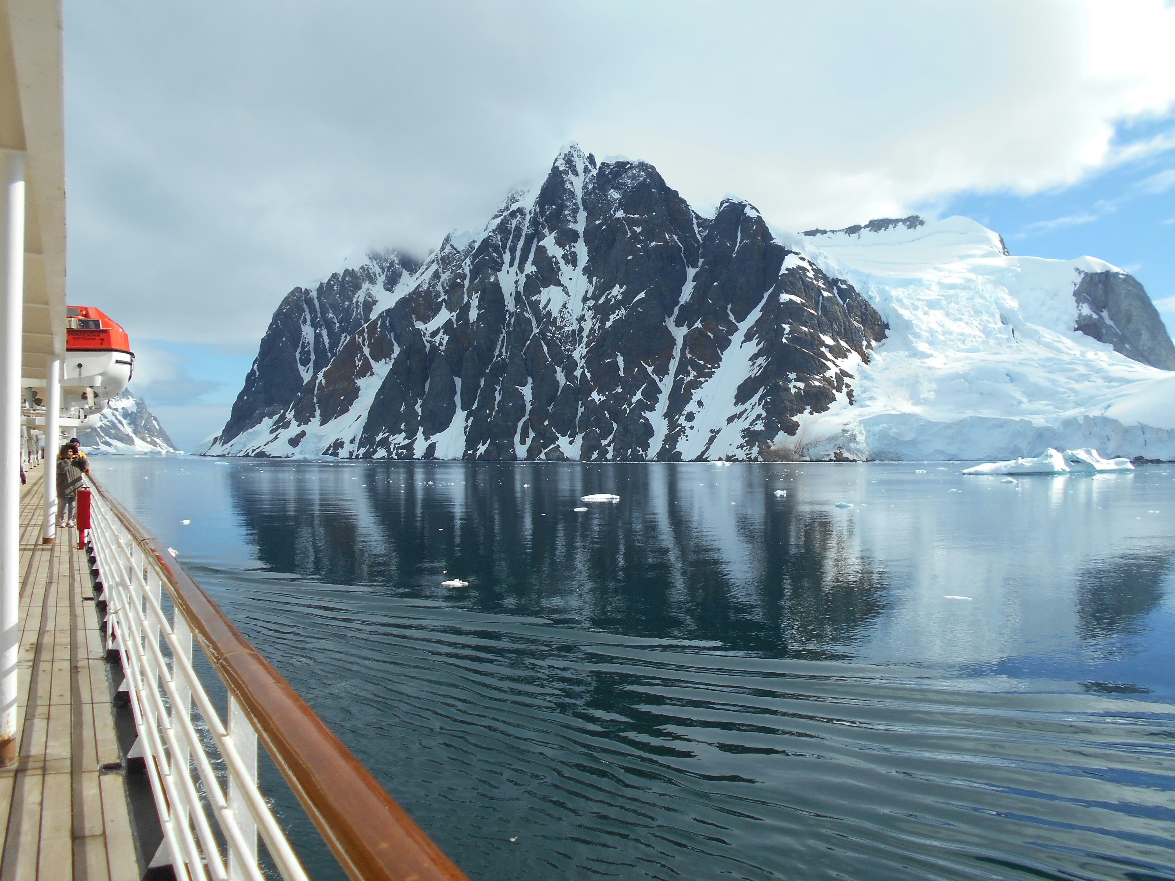 Snow-dusted mountain reflecting in calm blue water, viewed from a cruise ship's wooden deck.